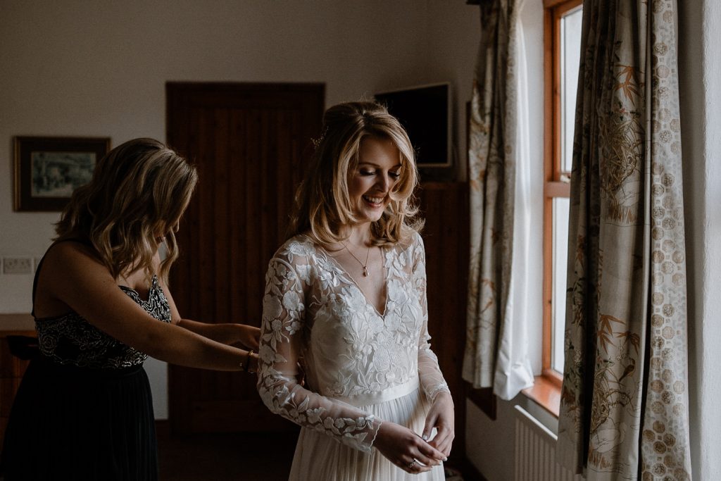 bride smiling as her sister buttons back of dress 
